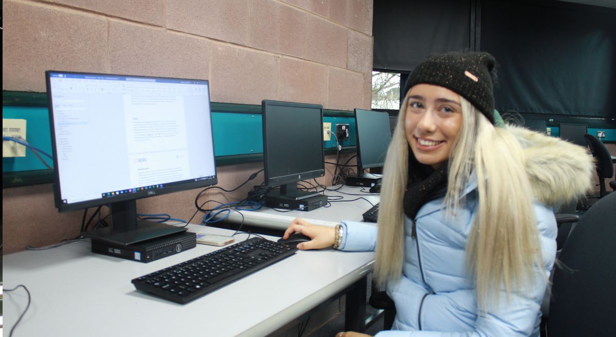 Female with long blonde hair, wearing a blue puffa jacket and black hat, smiling to camera and sitting at a computer desk.